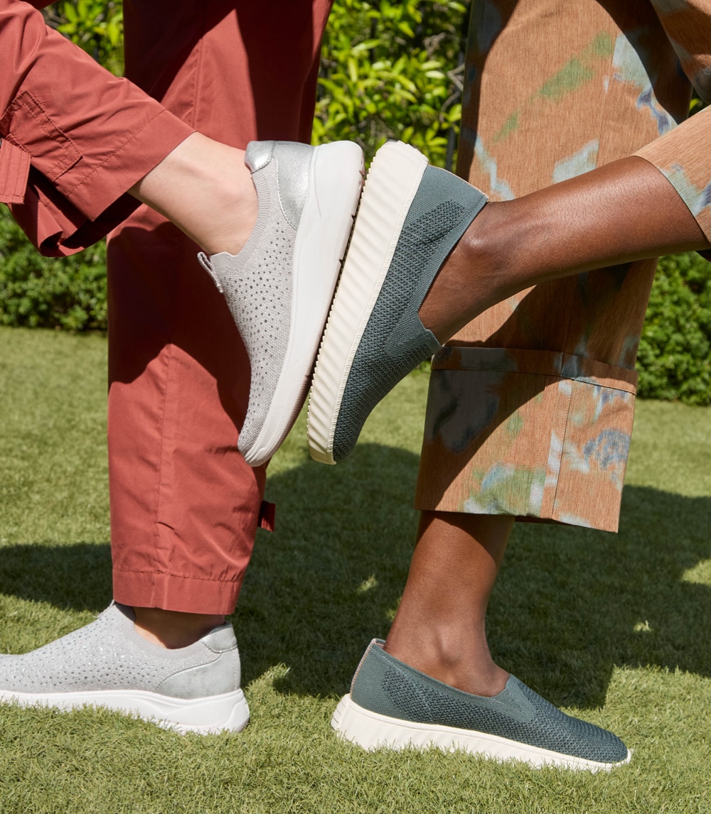 Two women standing back to back, each with one foot on the ground and the other foot lifted up and touching each other's shoe soles